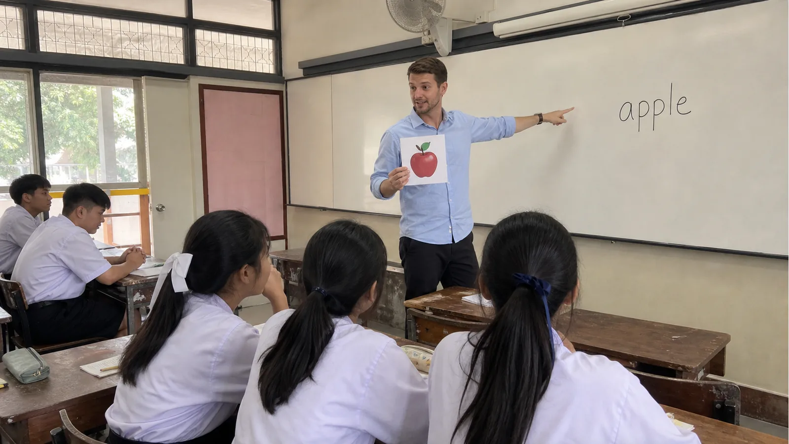 Foreign teacher pointing to the word apple on a whiteboard while holding an apple flashcard